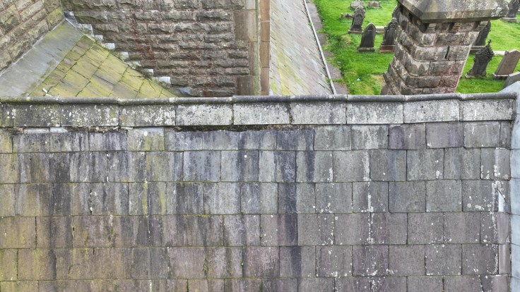 Aerial view of a slate roof with moss and lichen, leading to stone walls and nearby gravestones on a grassy area.